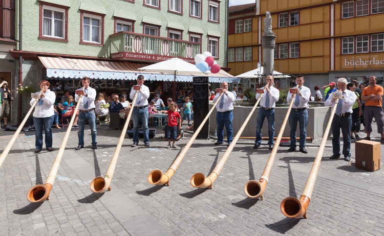 七位阿爾卑斯長號演奏者在小鎮上演奏。背景中，咖啡館的遊客正在聆聽音樂。